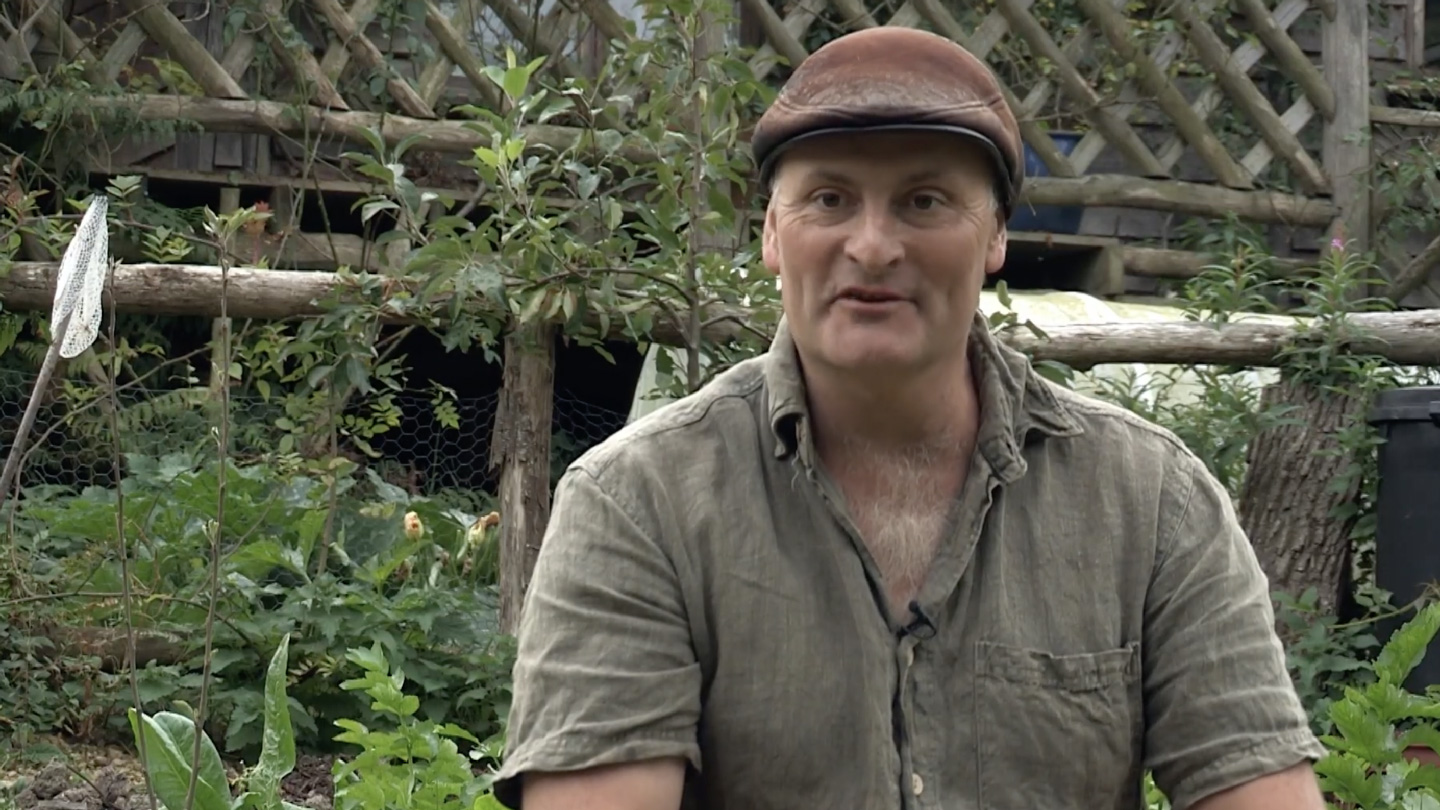Watch ‘Episode 1: This Off-Grid Dream Is Real!’ (Ben Law, seated, talking to camera in front of his self-built house. He wears a brown flat cap and green short-sleeve shirt.)